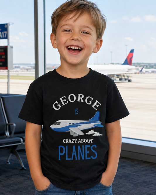A young boy smiling in an airport terminal wearing a black graphic t-shirt that reads 'GEORGE IS CRAZY ABOUT PLANES' with a blue and white airplane illustration.