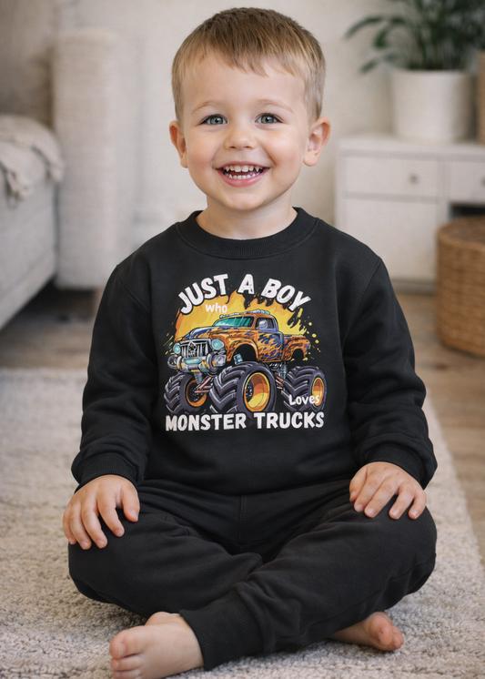 Smiling toddler sitting on rug wearing black “Just a Boy Who Loves Monster Trucks” sweatshirt and joggers, warm home lifestyle scene