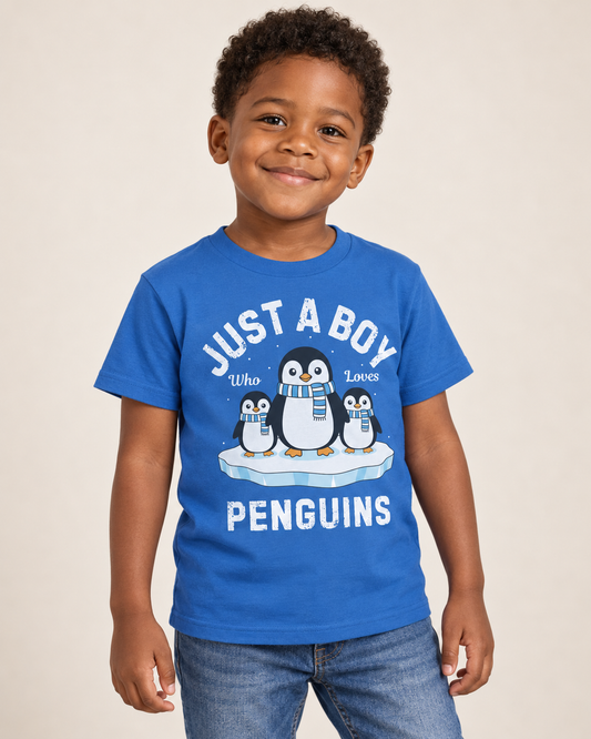 Young boy wearing a royal blue “Just a Boy Who Loves Penguins” T-shirt featuring three penguins in blue striped scarves on an ice floe, paired with jeans in a bright studio.
