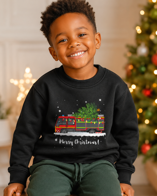 A smiling young boy wearing a black crewneck sweatshirt with a festive graphic of a red and green Christmas Fire Engine carrying a tree, with 'Merry Christmas!' written below. The background shows a softly blurred Christmas tree with warm lights.