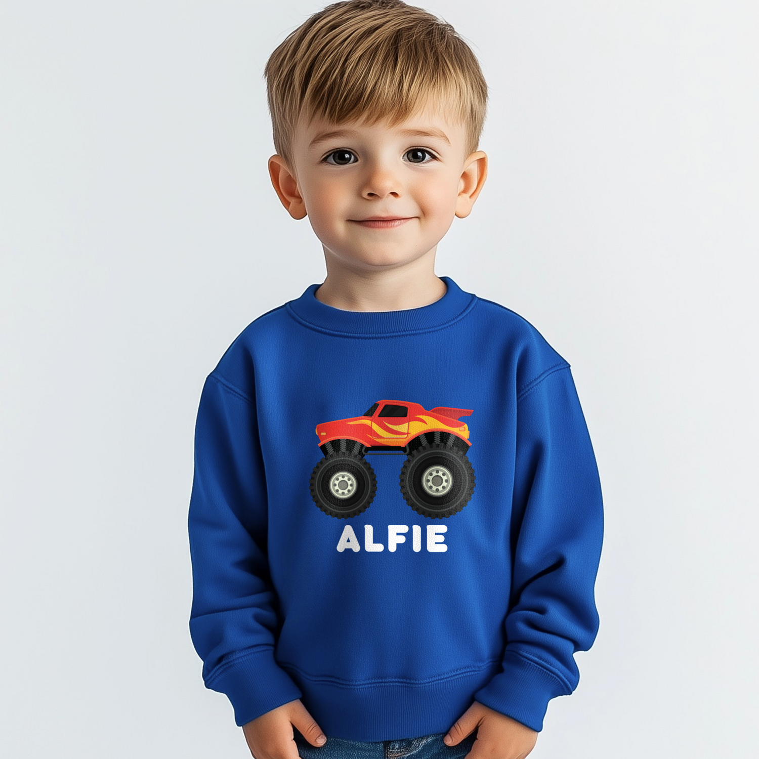 Young boy models a royal blue kids sweatshirt featuring a red and yellow monster truck graphic with large tyres and personalised with the name “Alfiel” printed below the design.