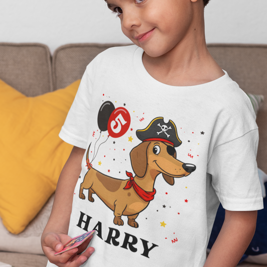 A young child models a white t-shirt. The shirt features a cartoon dachshund dressed as a pirate, wearing a hat and red bandana. Balloons with the number "5" are also part of the design, along with the name "HARRY" in black text. Small stars and confetti graphics are scattered around the main illustration.