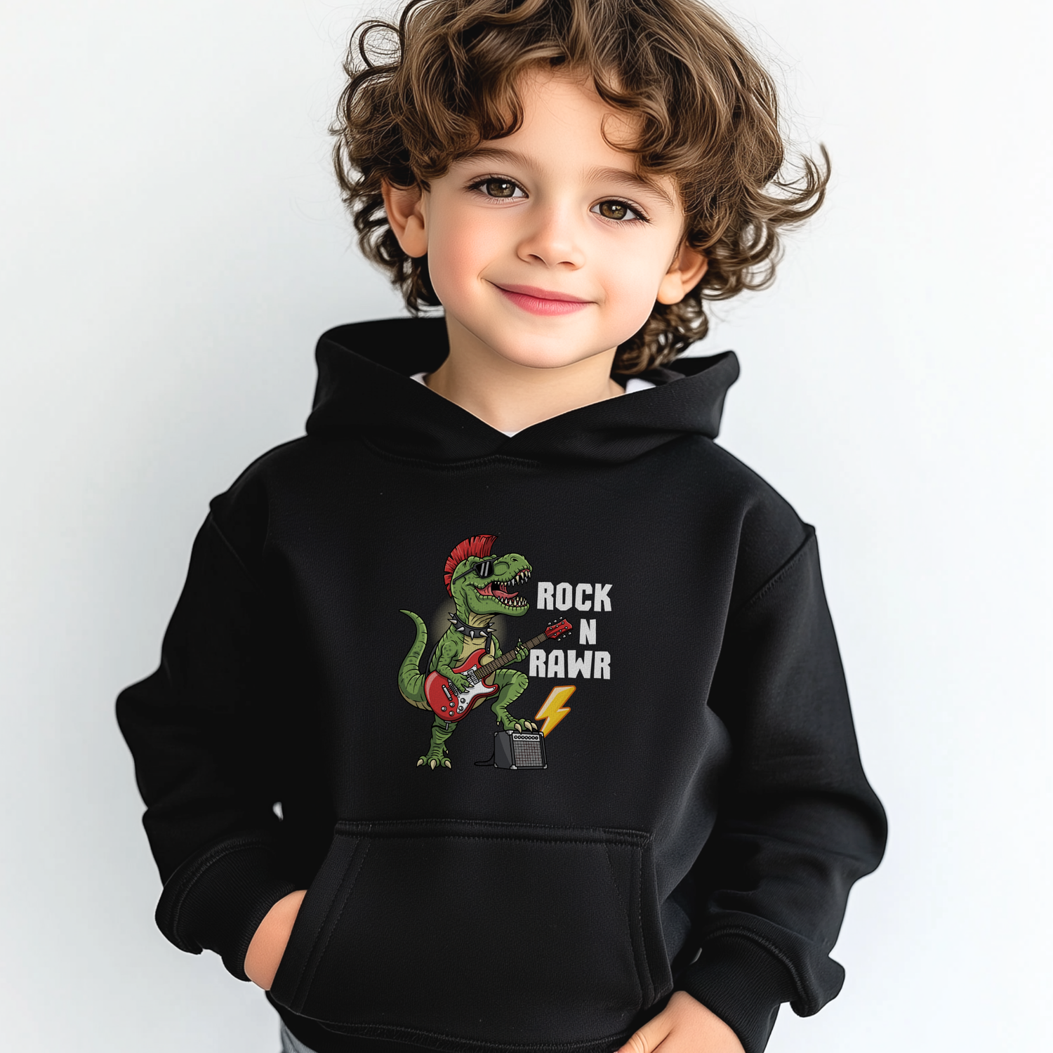 Young boy wearing a black “Rock N Rawr” dinosaur guitar hoodie, smiling with hands in pockets.