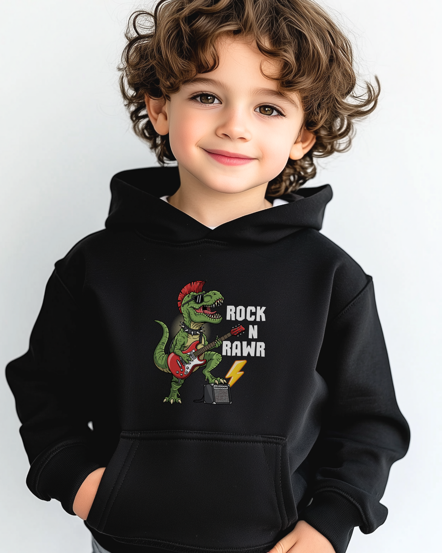Young boy wearing a black “Rock N Rawr” dinosaur guitar hoodie, smiling with hands in pockets.