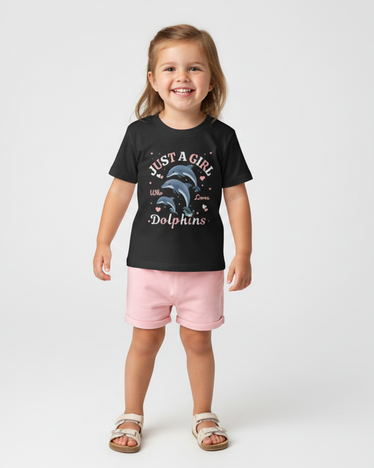 Little girl wearing black dolphin t-shirt with pastel pink shorts and sandals standing in studio