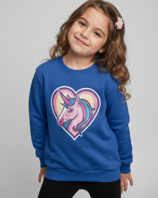 A young girl with wavy brown hair and a pink flower hair clip is wearing a royal blue crewneck sweatshirt with a colourful unicorn graphic inside a heart on the front, paired with black pants, against a solid light grey background.