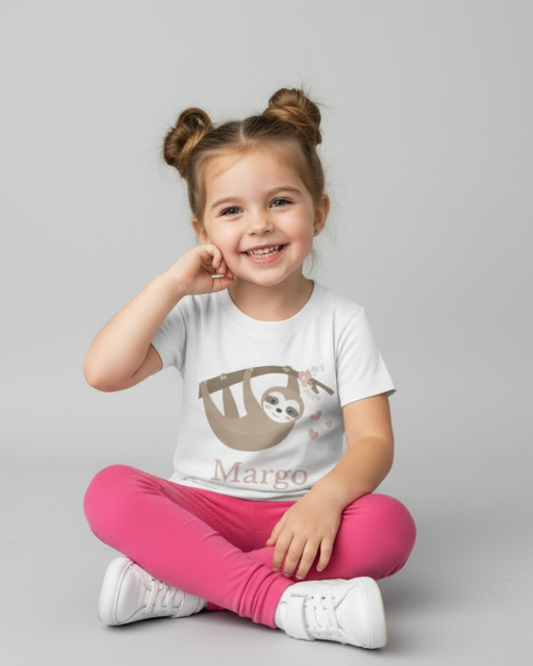 Smiling little girl sitting crossed legged wearing a white personalised kids t-shirt with a cute sloth illustration and the name “Margo”, studio grey background