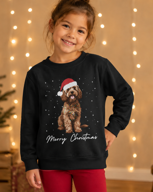 A young girl smiling while wearing a black crewneck sweatshirt with a graphic of a brown Cockapoo in a Santa hat and 'Merry Christmas' text, paired with red pants, set against a warm background of string lights.