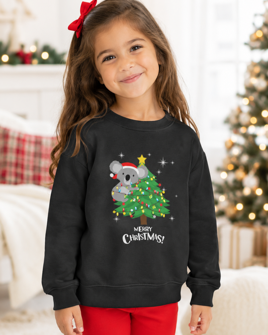 A young girl with a red bow in her hair is wearing a black crewneck sweatshirt with a festive Christmas koala and tree graphic, paired with red pants, set against a cozy, blurred holiday living room background.