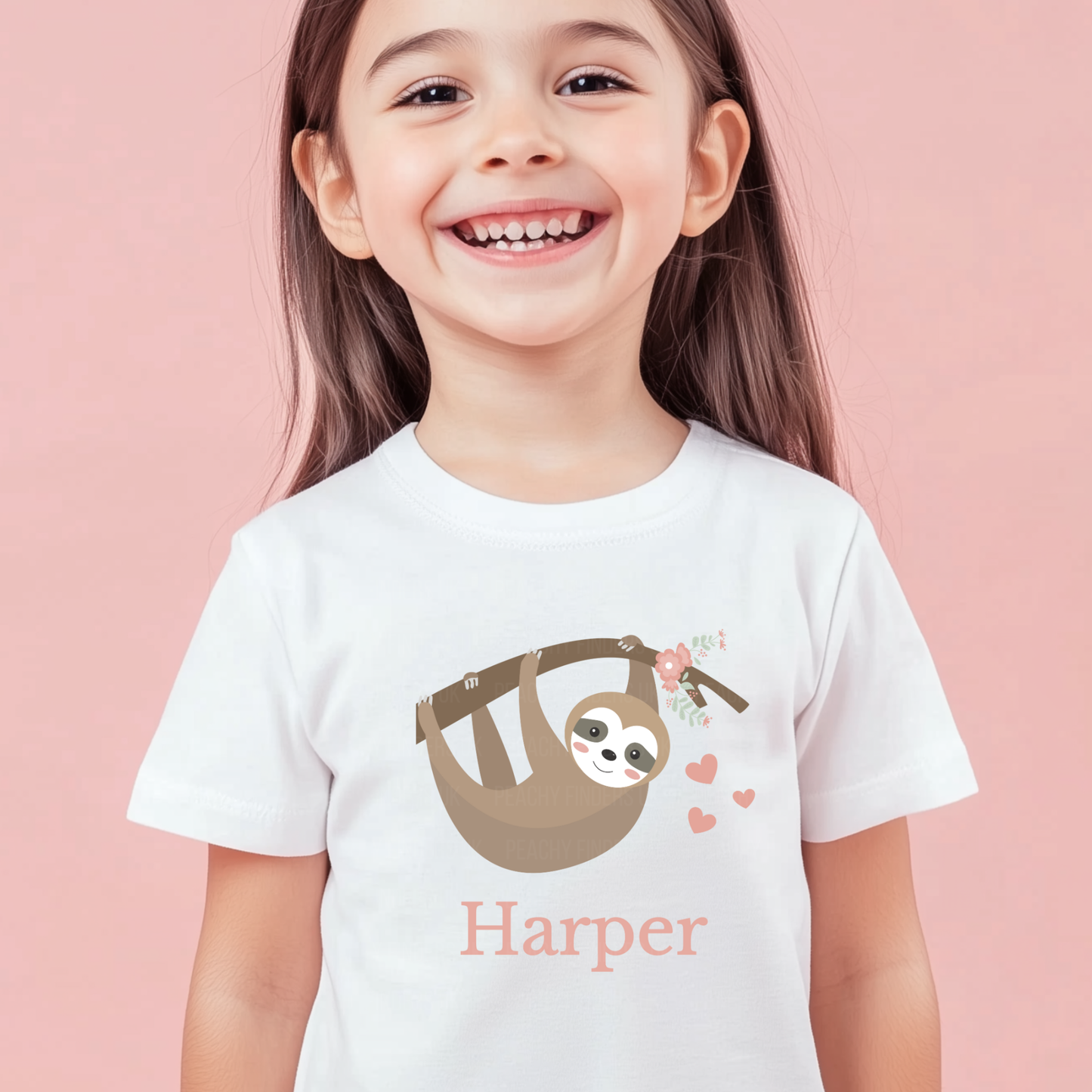 Smiling young girl wearing a white T-shirt with a cute hanging sloth, flowers and hearts above and the personalised name “Harper” printed underneath.