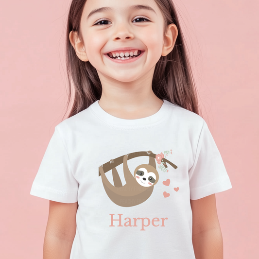 Smiling young girl wearing a white T-shirt with a cute hanging sloth, flowers and hearts above and the personalised name “Harper” printed underneath.