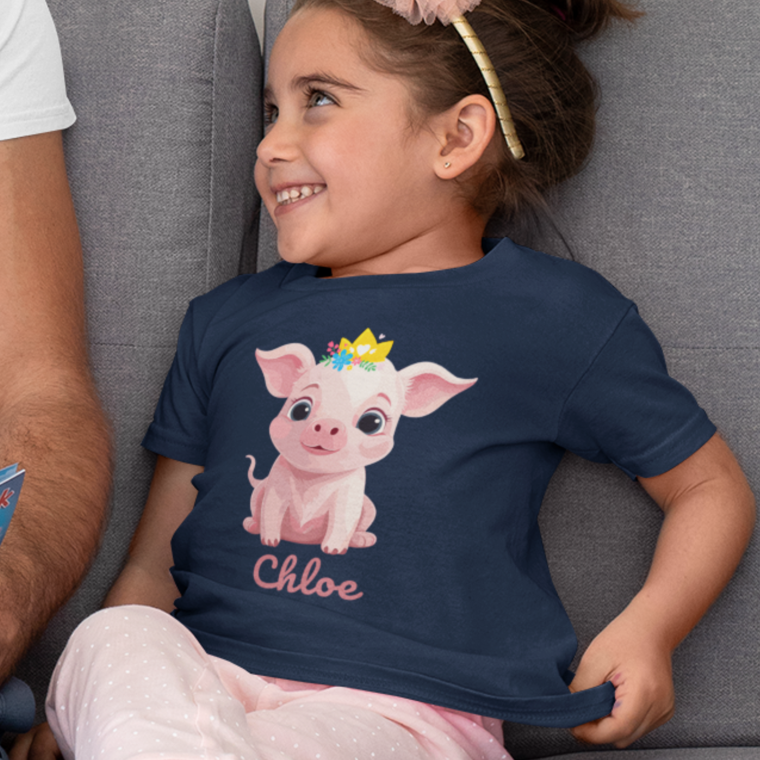 Smiling young girl sitting on a sofa wearing a navy blue t-shirt featuring a cute pink pig with a golden crown, along with the personalised name “Chloe” printed below the design.