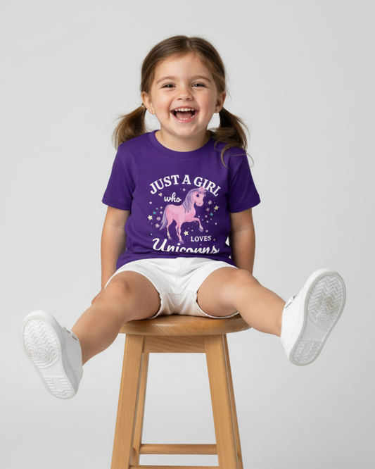 Happy young girl sitting on a wooden stool wearing a purple “Just a Girl Who Loves Unicorns” t-shirt with a pink unicorn design, white shorts, and white trainers, legs stretched out on a clean studio background.