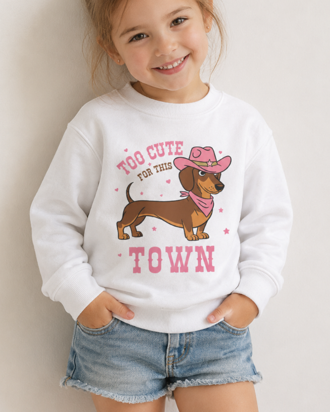 A young girl smiling and wearing a white crewneck sweatshirt with a graphic of a dachshund wearing a pink cowboy hat and bandana, with the text 'TOO CUTE FOR THIS TOWN' in pink. She is also wearing denim shorts against a plain light beige background.