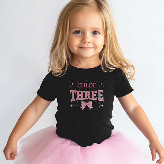 Young girl wearing a black personalised “Three” birthday T-shirt with pink bow graphic and a fluffy pink tutu, smiling against a plain light background.