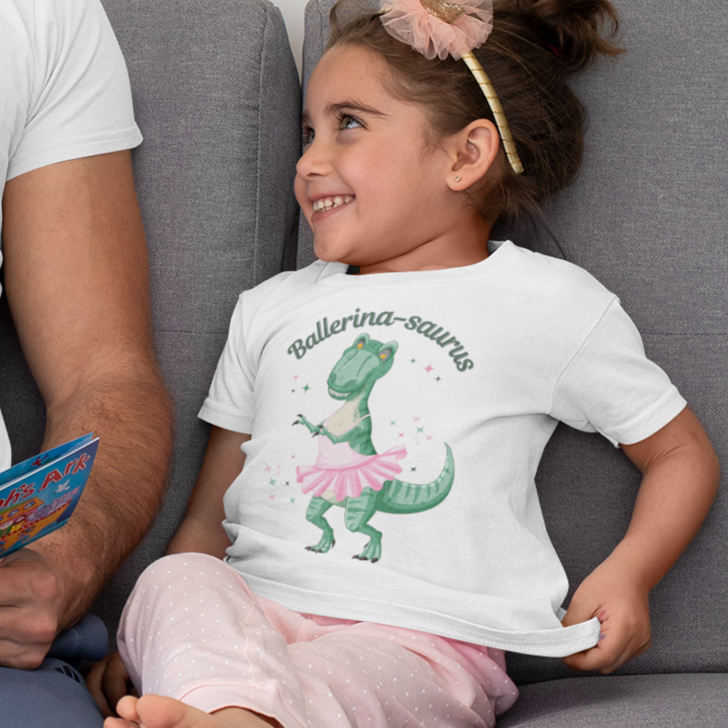 Little girl wearing a white Ballerina-saurus T-shirt with a green dinosaur in a pink ballet tutu, smiling on a sofa next to an adult.
