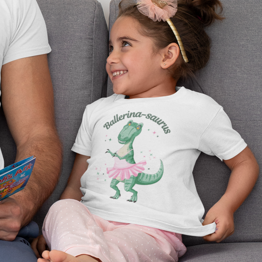 Little girl wearing a white Ballerina-saurus T-shirt with a green dinosaur in a pink ballet tutu, smiling on a sofa next to an adult.