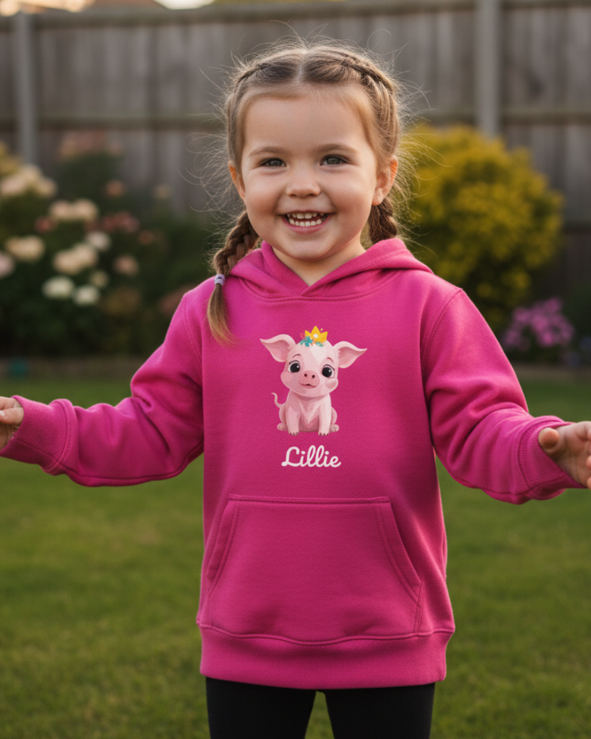 A young girl with braided hair stands outdoors on a grassy lawn wearing a bright pink hoodie with a cartoon pig graphic and the name 'Lillie' printed on the front, paired with black leggings.