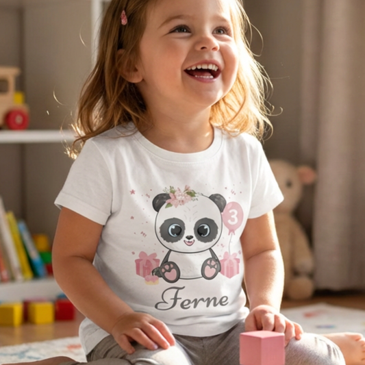 A little girl wearing a white t-shirt with a panda design and the name 'Ferne' in a room with toys and books.