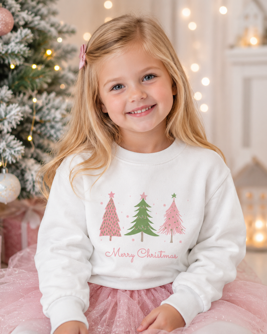 A young girl with blonde hair sits in front of a decorated Christmas tree, wearing a white crewneck sweatshirt with a pink and green Christmas tree graphic and 'Merry Christmas' text, paired with a pink tutu skirt.