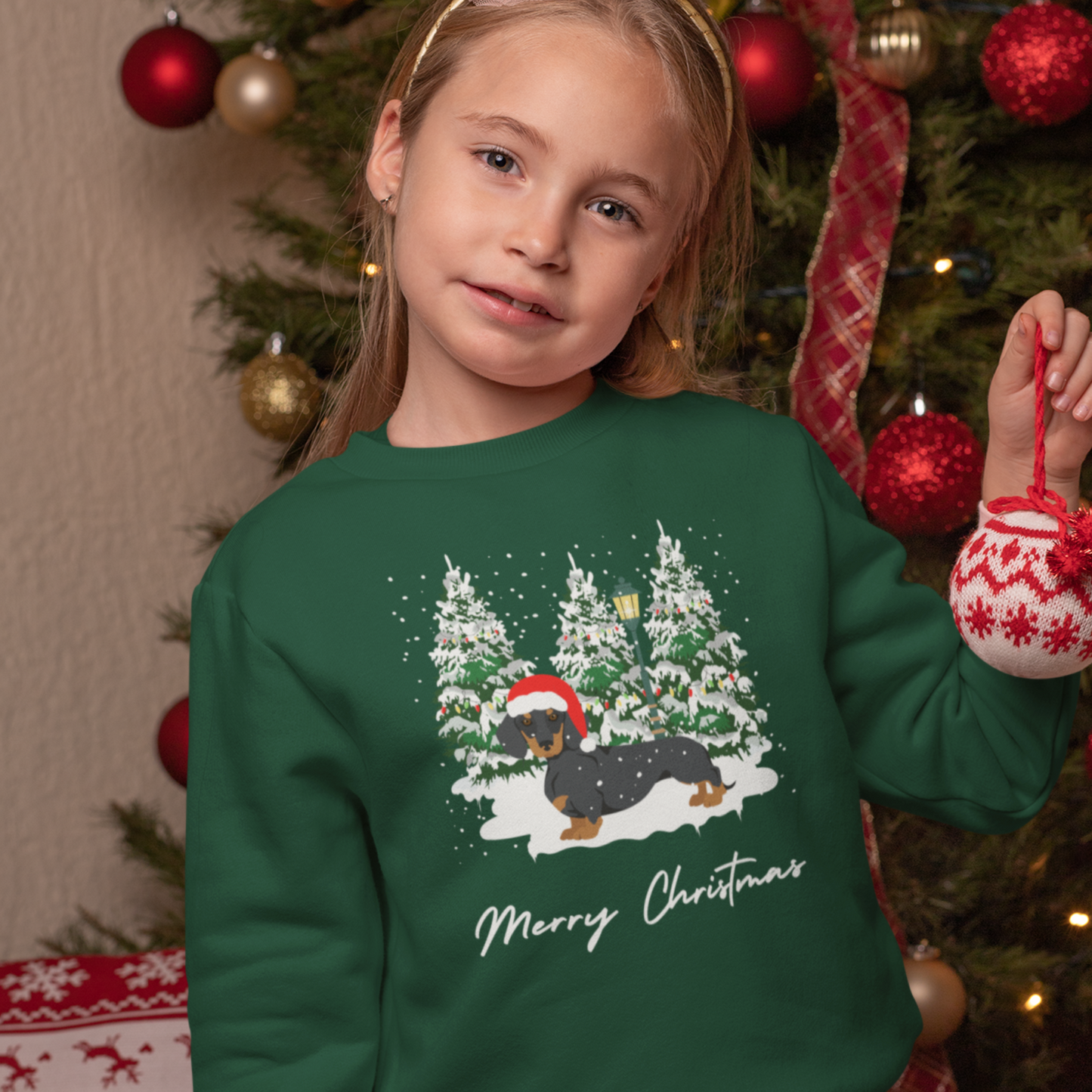 Young girl wearing a dark green Christmas sweatshirt featuring a festive dachshund in a Santa hat, surrounded by snowy Christmas trees and holding a red ornament near a decorated Christmas tree.