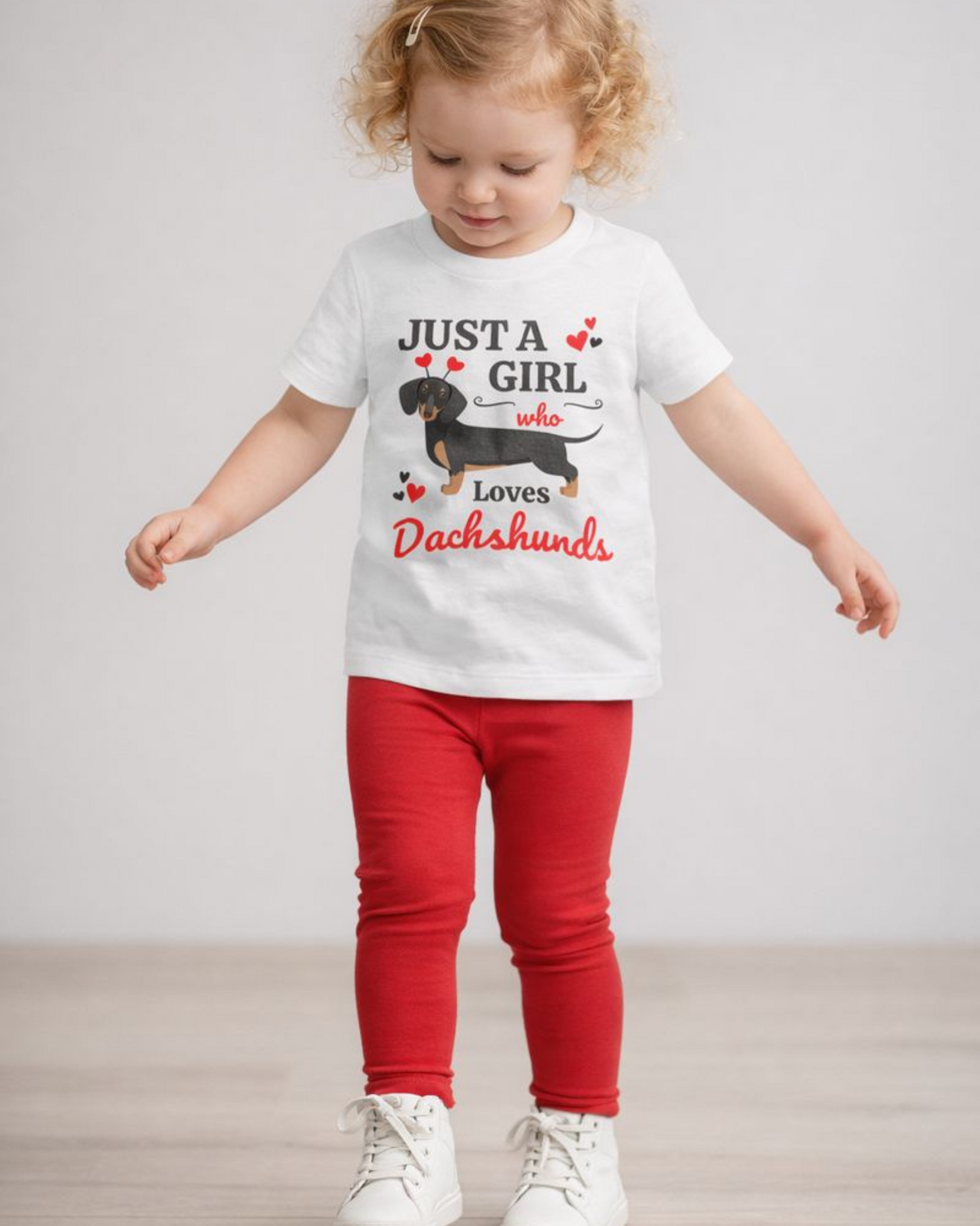 Blonde toddler girl wearing a white “Just A Girl Who Loves Dachshunds” t-shirt with red leggings and white trainers in a studio setting