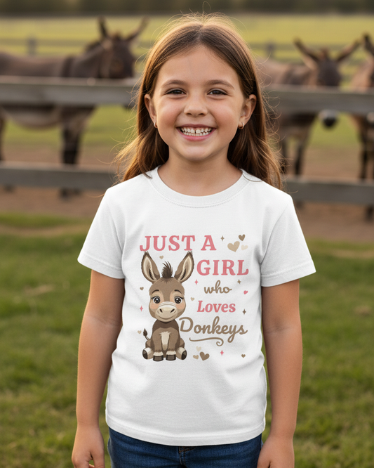 Smiling young girl wearing a white “Just a Girl Who Loves Donkeys” t-shirt standing in front of a wooden fence with donkeys in a grassy field, countryside lifestyle setting.