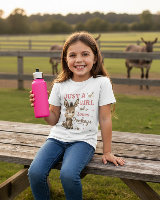 Happy young girl wearing a white “Just a Girl Who Loves Donkeys” t-shirt sitting on a wooden picnic table holding a pink water bottle, with donkeys grazing in a field behind her.