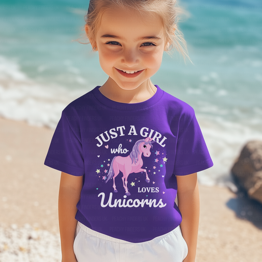 A young girl with light blonde hair pulled back smiles while wearing a vibrant purple t-shirt. The t-shirt features a graphic of a pink unicorn with a rainbow mane and tail, surrounded by stars and the text "JUST A GIRL who Loves Unicorns". The background is a bright, blurred beach scene with light sand and turquoise sea.