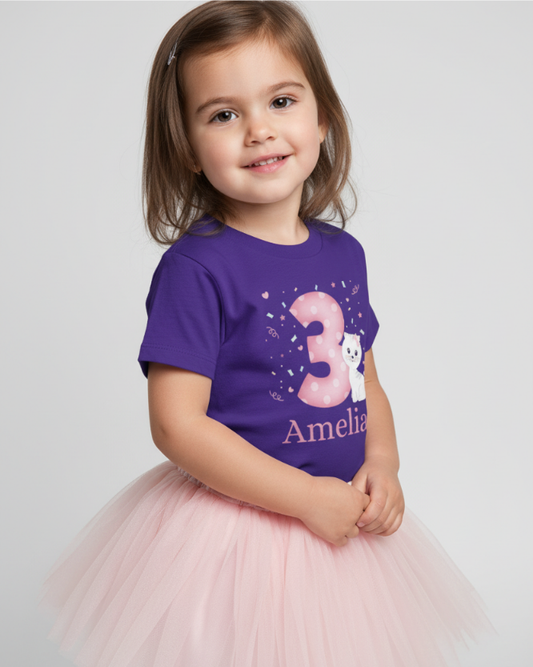 Smiling young girl modelling a personalised purple third birthday shirt with pink polka dot number 3 and kitten graphic, name Amelia, paired with a fluffy pink tutu in a bright studio setting