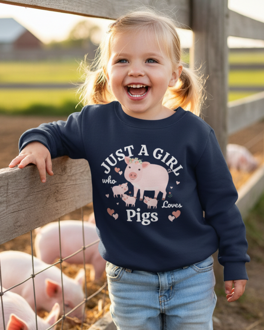 A young child wearing a navy blue sweatshirt with a pig graphic and the text 'JUST A GIRL who loves Pigs' stands smiling next to a wooden fence with pigs in a farm pen.