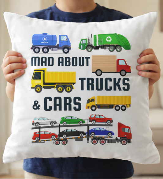 Close-up of a child holding a “Mad About Trucks & Cars” cushion with bright illustrated trucks, lorries, and cars on a white pillow.
