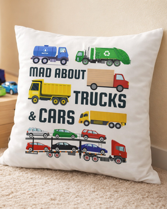 Kids decorative cushion with “Mad About Trucks & Cars” design featuring colourful cartoon trucks and cars, styled in a child’s bedroom floor with toy vehicles in the background.
