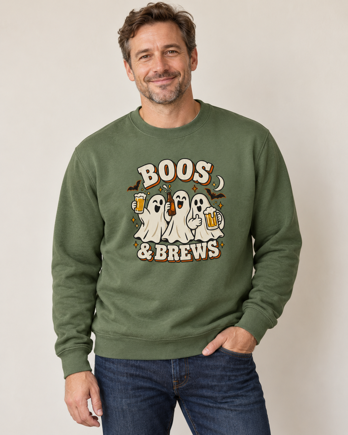 A man wearing an earthy green crewneck sweatshirt with a 'Boos & Brews' Halloween graphic featuring ghosts and beer mugs, paired with blue jeans in a studio setting.