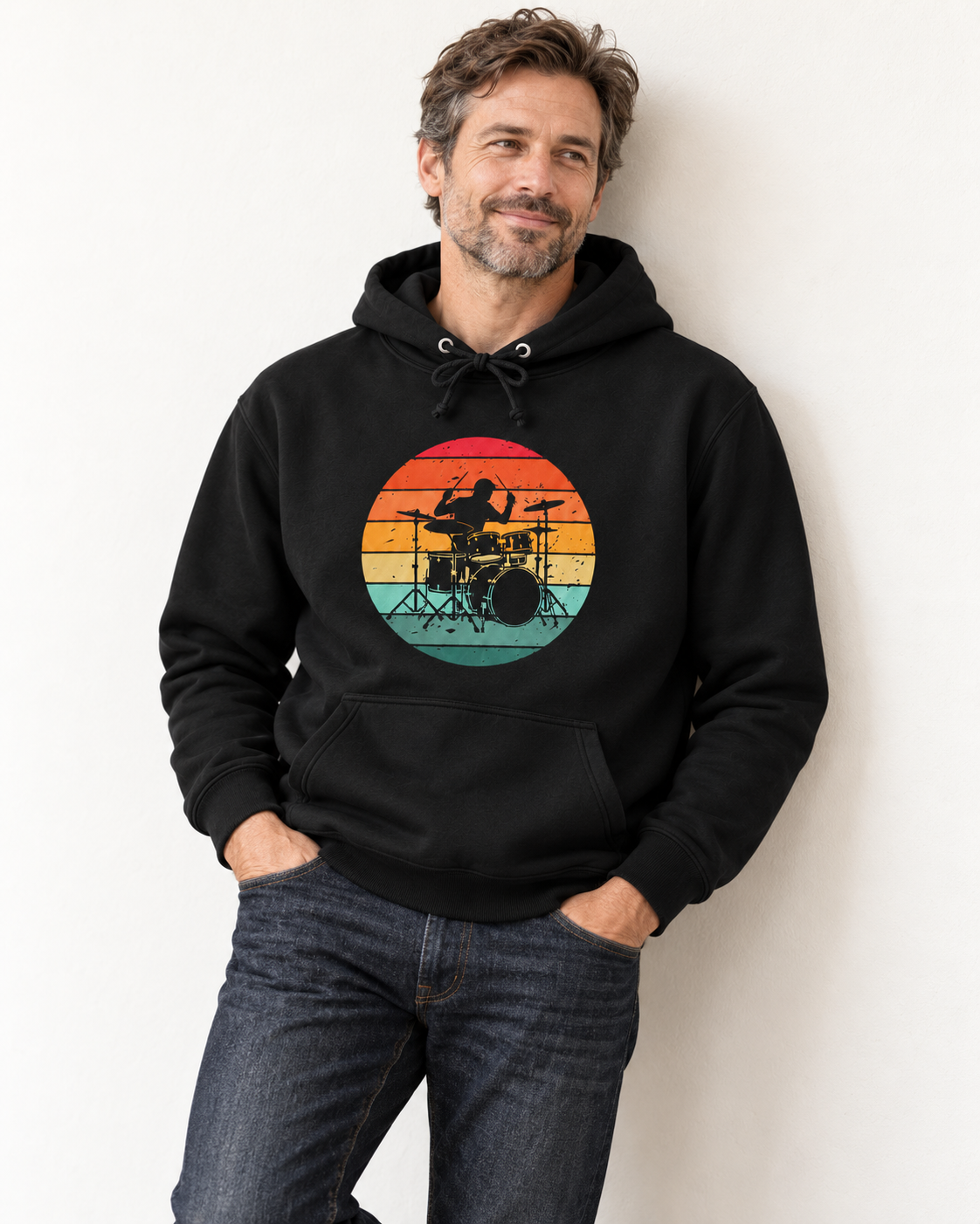 A man wearing a black pullover hoodie with a circular retro sunset graphic of a drummer, paired with dark blue jeans, standing against a plain white wall.
