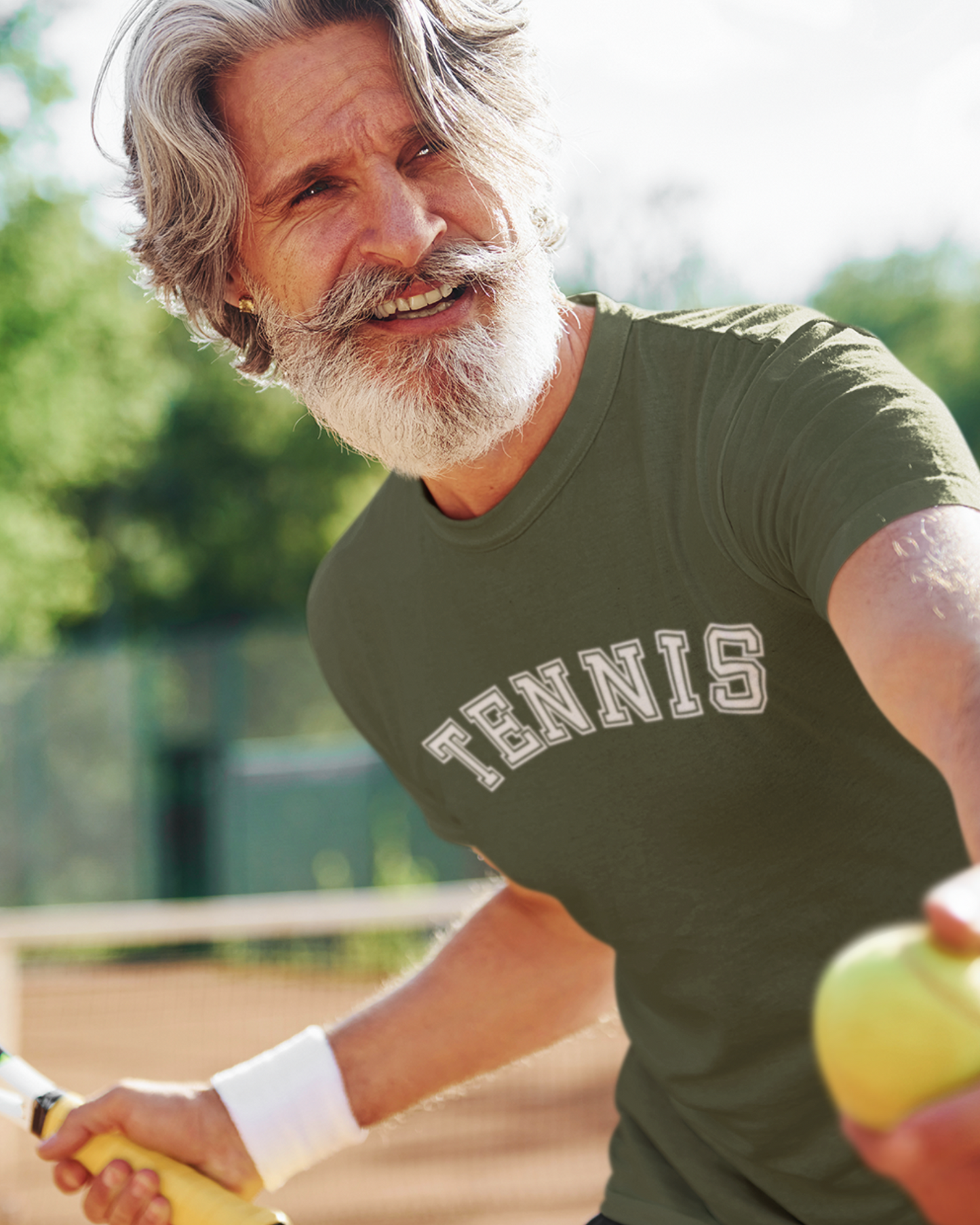 Man on a tennis court wearing a military green Tennis slogan T-shirt and holding a racket and ball.
