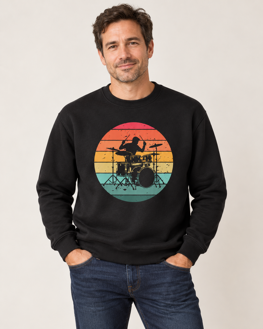 Man wearing a black retro drummer sweatshirt with blue jeans, standing with his hands in his pockets against a light studio background.
