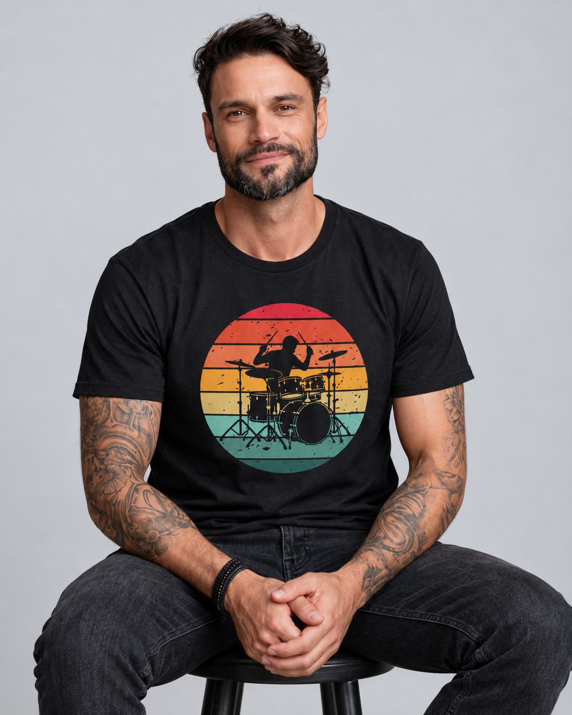 Man seated on a black stool wearing a black retro drummer T-shirt with dark jeans, with the sunset drum kit graphic fully visible.