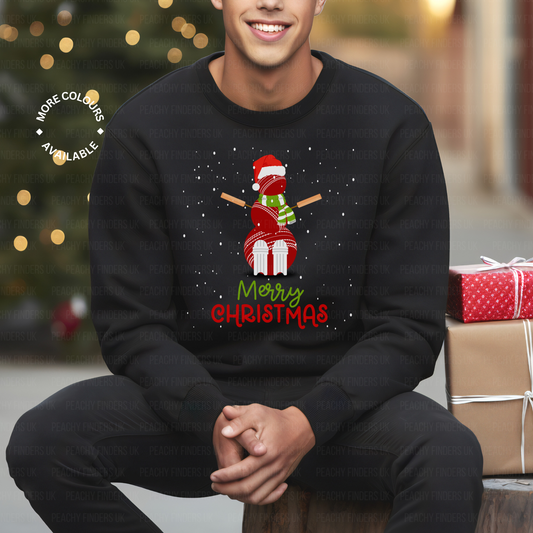 Smiling man wearing a black Christmas jumper featuring a festive snowman made from red cricket balls, stumps, and bats, with "Merry Christmas" text, sitting next to wrapped presents and a lit Christmas tree in the background.