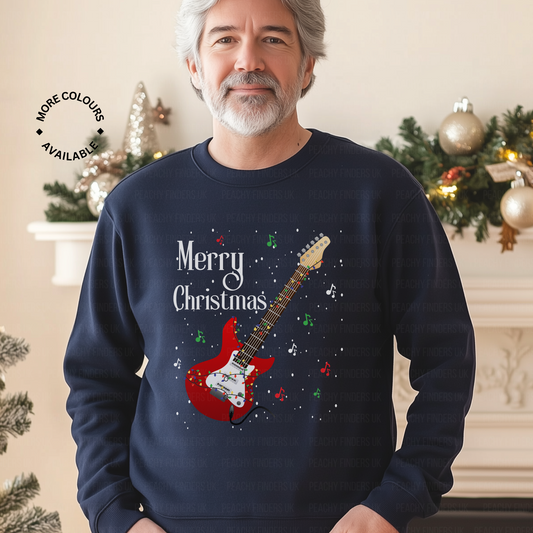Man wearing a navy blue Christmas sweatshirt with a red guitar wrapped in fairy lights and musical notes design with 'Merry Christmas' text, standing in a festive decorated room.