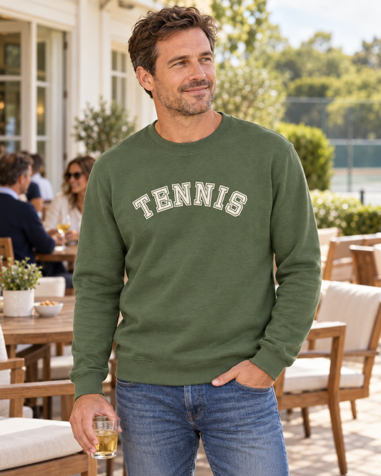 Man wearing an earthy green varsity tennis sweatshirt holding a drink on an outdoor clubhouse terrace.