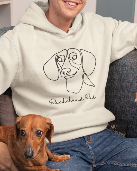 A man wearing a cream-coloured hoodie with a black line art graphic of a dachshund and the text 'Dachshund Dad' sits on a couch with a brown dachshund dog on his lap.