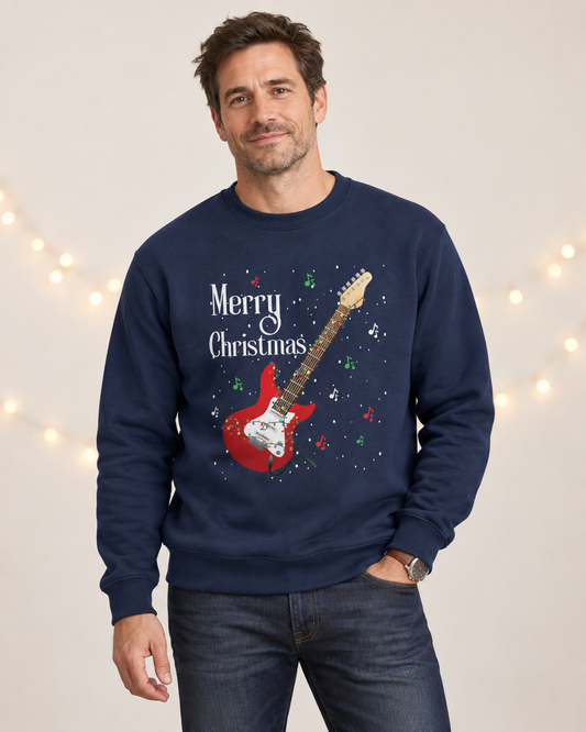 A man wearing a navy blue crewneck sweatshirt with a 'Merry Christmas' and red electric guitar graphic, paired with dark blue jeans, set against a warm beige background with soft string lights.