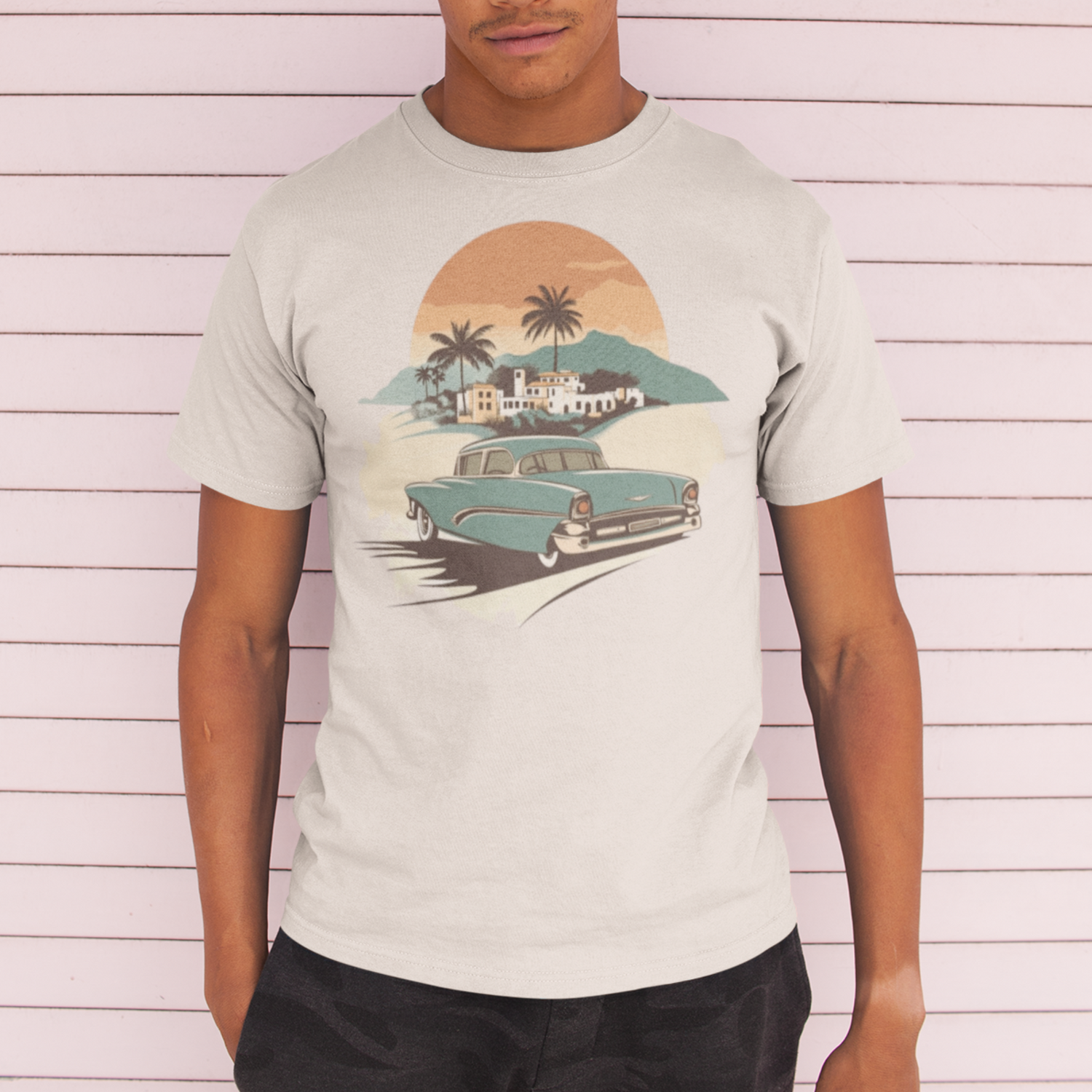Man in vintage car t-shirt with sunset, palms, and town graphic. Wearing black trousers, against a pink and white wood panel background.