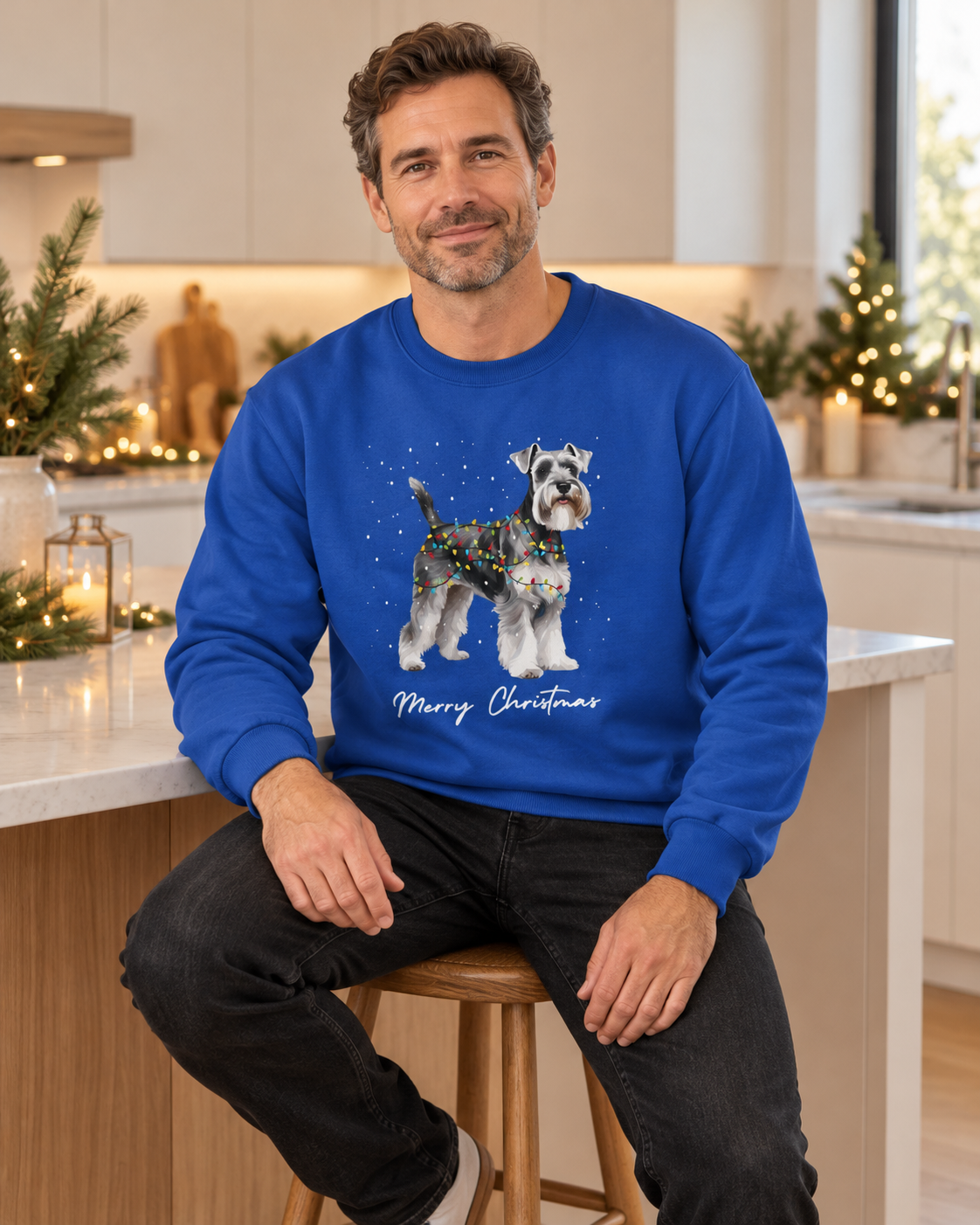 Man wearing the royal blue Christmas Schnauzer jumper sitting on a stool in a festively decorated modern kitchen