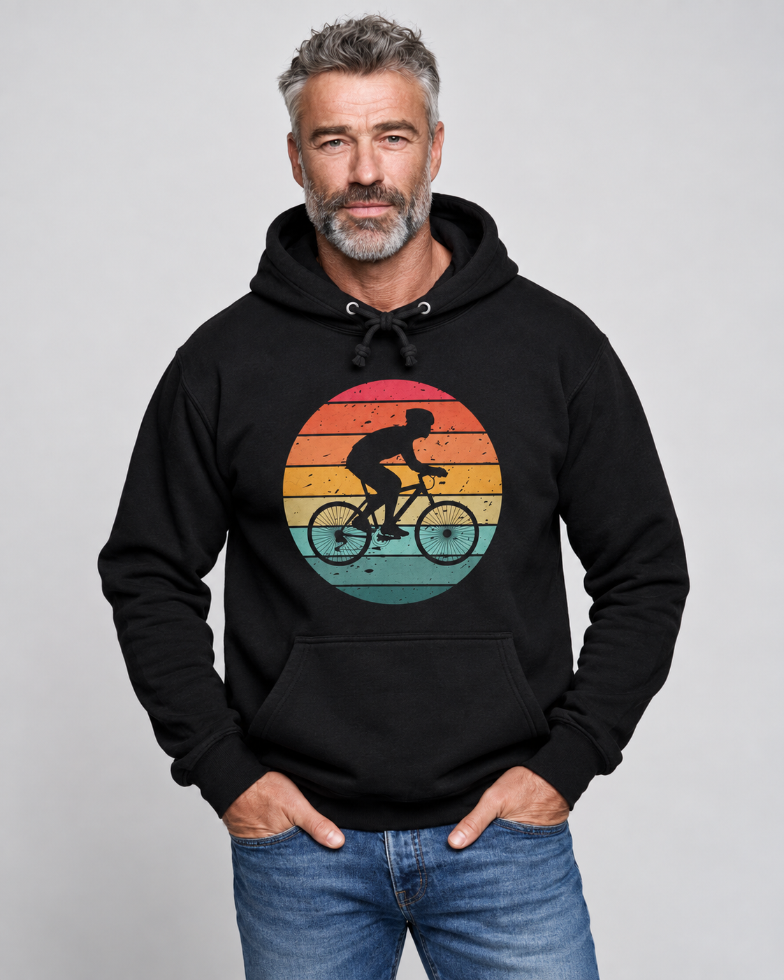 A man wearing a black pullover hoodie with a retro circular graphic of a cyclist against a sunset gradient, paired with blue jeans, photographed against a neutral gray background.