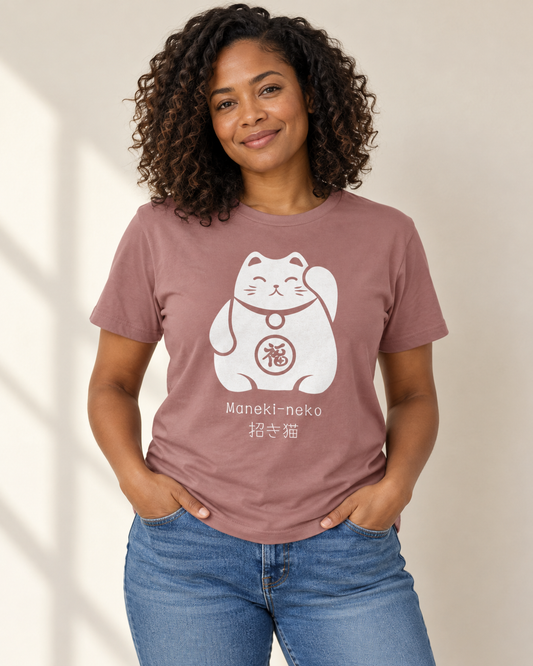 A woman with curly dark hair wearing a mauve pink short-sleeve t-shirt and blue jeans, standing against a neutral beige background. The t-shirt features a white illustration of a Japanese lucky cat with the words "Maneki-neko" and Japanese kanji characters printed below