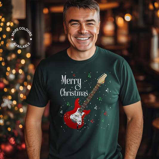 A man smiling, wearing a dark green navy t-shirt with a 'Merry Christmas' text and a graphic of an electric guitar, standing in a room with Christmas decorations.