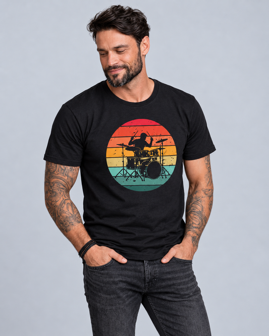 Man wearing a black sunset drummer T-shirt and dark jeans, standing with his hands in his pockets and looking down to the side.