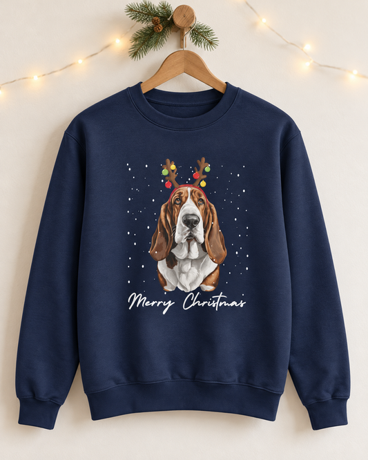 A navy blue crewneck sweatshirt hanging on a wooden hanger against a warm off-white wall. The sweatshirt features a graphic of a Basset Hound wearing Christmas reindeer antlers, surrounded by falling snow, with 'Merry Christmas' written in white script below.